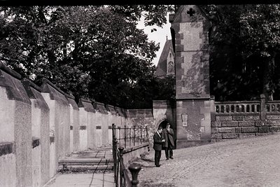 Black-and-white street scene featuring a stone wall cemetery with arched niches and a central tower. Two figures walk along a...