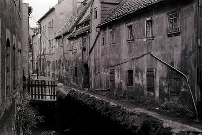 Narrow, cobblestone alley flanked by aged, plastered buildings with wooden-framed windows—likely Eastern European. Stone brid...