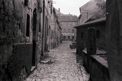 Narrow cobblestone alleyway flanked by aged stone buildings, likely European. Signage in German ("Café") suggests a mid-20th ...