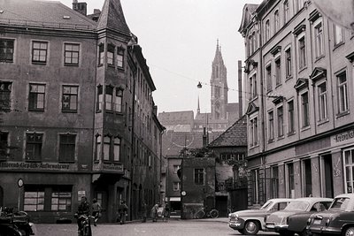 Mid-20th century European street scene with classic brick buildings and vintage signage. Prominent church spire in background...