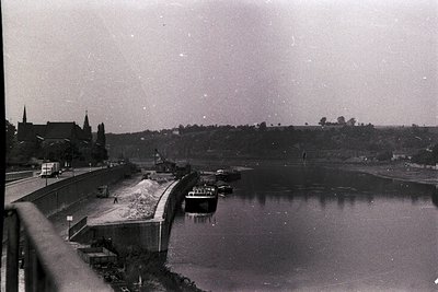 Vintage black-and-white shot of a river with a concrete dam and docked barge, likely mid-20th century. Distant church steeple...