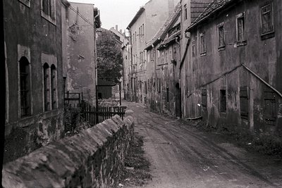 Narrow, unpaved alley flanked by aged stone buildings with arched windows and shuttered doors. Overgrown vegetation and a low...