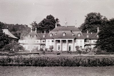 Neoclassical mansion with symmetrical façade, central portico, and mansard roofs—likely early-to-mid 20th century. Lush hedge...