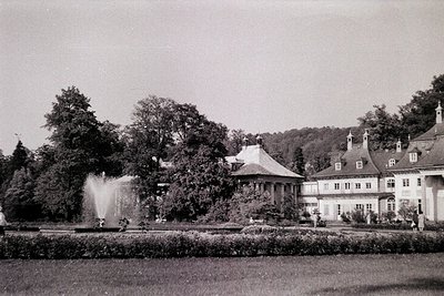 Neoclassical estate with symmetrical façade, gabled roofs, and decorative dormers. Central fountain sprays water into a manic...