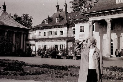 Neoclassical mansion with symmetrical façade, tall columns, and mansard roof in mid-20th century. Woman in 1960s-style coat p...