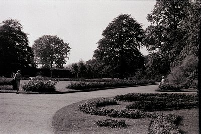 Symmetrical formal garden with manicured hedges and gravel paths, likely from the 1950s–1970s. Two figures—one standing, one ...