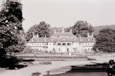 Neoclassical mansion with symmetrical façade, tall windows, and red-tiled roof, framed by manicured gardens and dense forest....