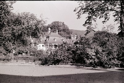 Vintage sepia-toned photo of a two-story estate with steep gabled roofs, surrounded by dense foliage. Central chimney and dor...