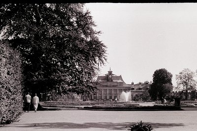 Neoclassical palace framed by symmetrical gardens, featuring a central fountain and manicured hedges. Two figures walk under ...