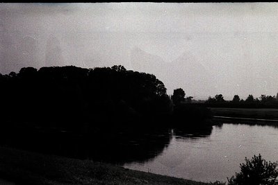 Vintage black-and-white landscape featuring a serene river bordered by dense tree foliage on both sides. The composition high...