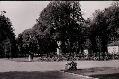 Vintage black-and-white park scene featuring a central classical urn monument on a pedestal, surrounded by manicured shrubs a...