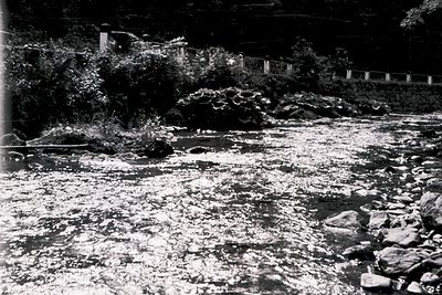 Black-and-white photo of a rocky riverbed with shallow, fast-moving water. Stone retaining walls and a low stone fence frame ...