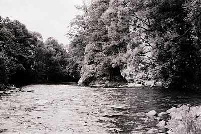Black-and-white river scene featuring a rocky gorge with a natural arch framing dense forest. Water flows swiftly over smooth...