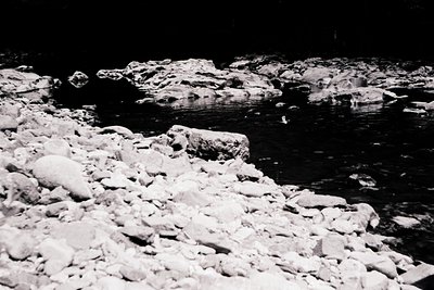 High-contrast black-and-white shot of jagged river rocks and shallow water at night, illuminated by faint ambient light. The ...
