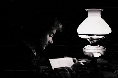 Vintage black-and-white portrait of an individual reading by a kerosene lamp, likely mid-20th century. The lamp’s glass shade...