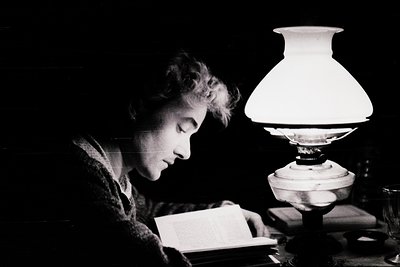 Black-and-white portrait of an individual engrossed in reading by kerosene lamp light, mid-20th century. Classic vintage lamp...