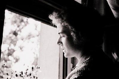 Black-and-white portrait of an individual gazing out a window at blurred foliage, suggesting contemplation or introspection. ...