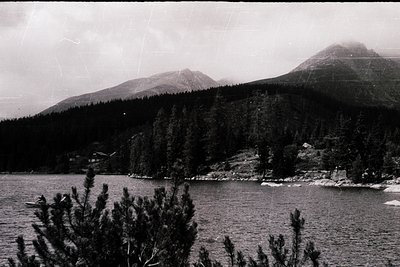 Black-and-white alpine lake scene with rugged coastline and dense coniferous forest. Snow-capped peaks rise in the background...