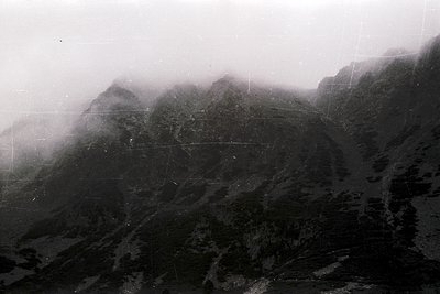 Vintage black-and-white aerial shot of rugged mountain terrain shrouded in mist, revealing jagged peaks and winding paths. Li...