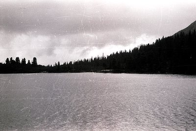 Frozen alpine lake bordered by dense coniferous forest under overcast skies. Ice texture suggests winter conditions. Vintage ...