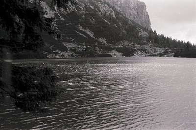 Black-and-white shot of rugged alpine lake nestled between sheer rock cliffs and dense coniferous forest. Water reflects ligh...