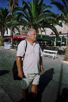 Mid-20th century man in a checkered shirt and shorts strolls past palm-lined promenade with stone buildings. Red Pepsi sign a...
