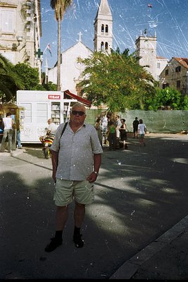 Mid-20th century street scene featuring a man in a checkered shirt and shorts standing under confetti. Behind him, a white ch...