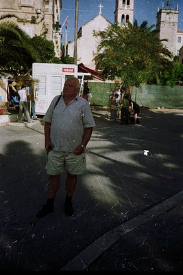Man in 1970s Mediterranean streetwear—striped polo, khaki shorts, and dark shoes—poses confidently in a sunlit plaza. Whitewa...