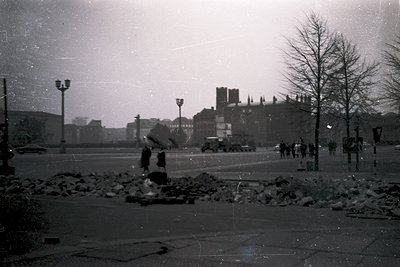 Mid-20th century urban scene showing post-war devastation: barricades of rubble block a street, a lone figure in dark clothin...