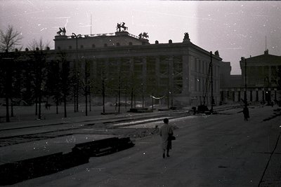 Neoclassical building with sculptural reliefs and columns dominates a wide, tree-lined boulevard. Mid-century urban scene sho...