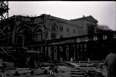 War-damaged neoclassical building facade with arched windows and columns, partially collapsed. Signage reads "UNSE" (likely p...
