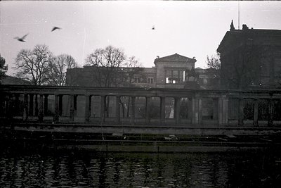 Neoclassical building reflected in calm water, framed by stone bridge railing. Overcast sky with birds in flight. Architectur...
