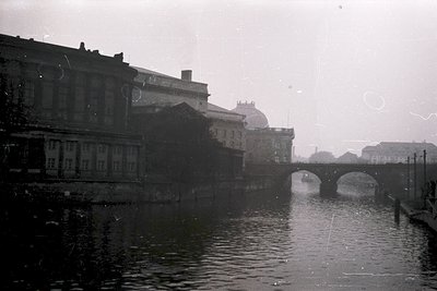 Vintage black-and-white canal scene featuring a stone bridge with three arches. Surrounding buildings exhibit early 20th-cent...