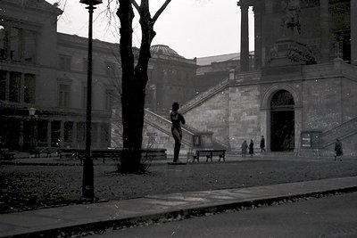 Neoclassical courtyard with grand stone columns and arched entryway. A lone figure in mid-stride descends wide stone stairs, ...