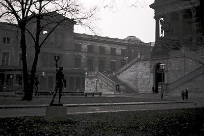 Neoclassical building with grand staircase and dome in background, flanked by leafless trees. Prominent statue of a figure in...