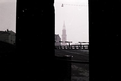 Black-and-white urban scene framed by a window sill, showing a church steeple and low-rise buildings. Likely mid-20th century...
