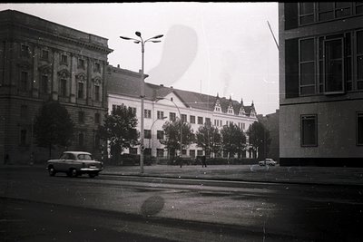 Mid-20th century urban street scene featuring classic European architecture. Prominent multi-story building with steep gables...