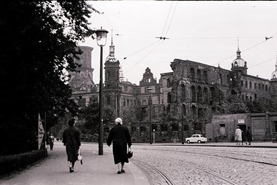 Black-and-white street scene featuring two women walking on a cobblestone path beside tram tracks. Prominent Soviet-era archi...