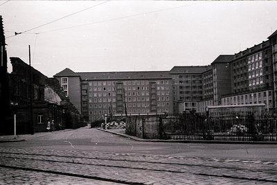 Mid-20th century urban street with Soviet-era Brutalist apartment blocks flanking a wide, empty road. Concrete facades, barre...