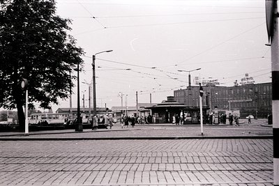 Black-and-white street scene featuring a tram stop with overhead wires and vintage tram tracks. Crowds gather near a central ...
