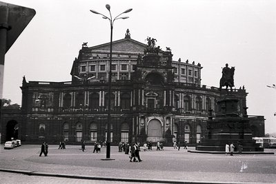 Grand neoclassical opera house with monumental columns, sculpted eagles, and Latin inscriptions. Pedestrians and vintage cars...