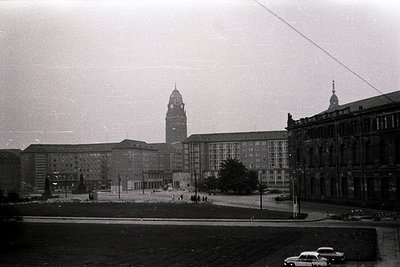 Mid-century Brutalist-style train station with prominent clock tower, flanked by long, rectangular concrete blocks. Empty pla...