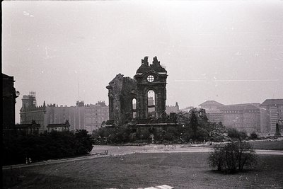 Ruins of a Gothic-style triumphal arch in a bombed urban landscape, mid-20th century. Surrounding buildings show war damage, ...