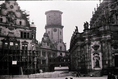 Historic European plaza featuring a grand Baroque-style tower flanked by ornate facades. Scaffolding indicates restoration wo...