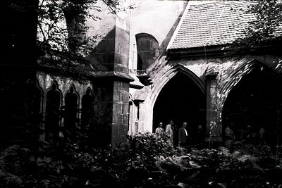 Gothic-style stone archway with pointed arches and buttresses, partially obscured by foliage. Group of people in 19th-century...