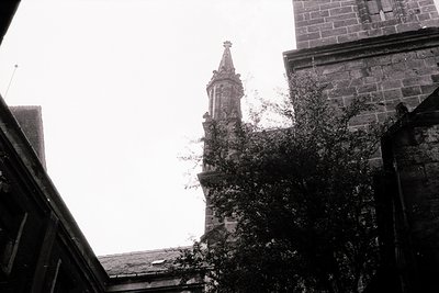 Gothic-style stone bell tower with intricate tracery, framed by dense foliage. Architectural details include a cross-topped s...