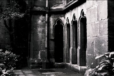 Gothic-style stone cloister corridor with pointed arch windows, weathered stonework, and low-angle lighting. Likely medieval ...