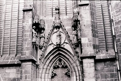 Gothic stone façade featuring intricate carvings: three statues flanking a pointed arch doorway, central shield emblem, and d...