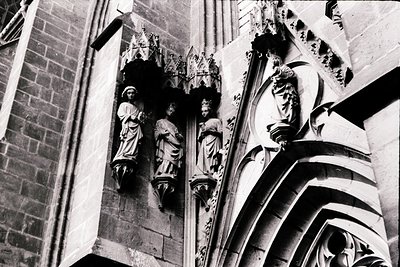 Gothic stone carvings depicting religious figures in niches, likely part of a cathedral façade. Intricate tracery and pointed...