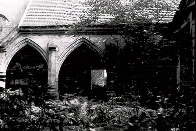 Gothic-style arched doorway with stone detailing, partially obscured by overgrown foliage. Interior reveals exposed brick and...